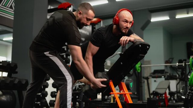 The coach monitors the correctness of the student's exercise with dumbbells in the gym. Bodybuilding classes on special equipment.