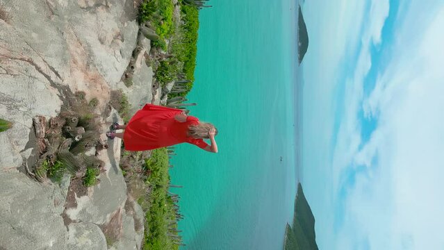 Woman on red dress standing on the coast with crystalline turquoise sea in Arraial do Cabo, Rio de janeiro, Brazil. Aerial drone vertical view.