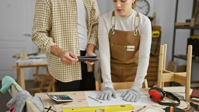A man and woman collaborate in a well-equipped carpentry workshop, analyzing designs and using various woodworking tools.