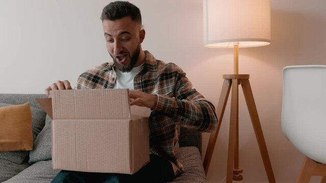 A smiling young man excitedly opening a delivery box at home, representing a successful online shopping experience.