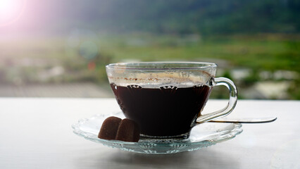 A cup of hot black coffee with brown sugar on plate on white table and blur green nature outdoor background. Morning coffee concept. Good start for morning inspiration and fresh energy.