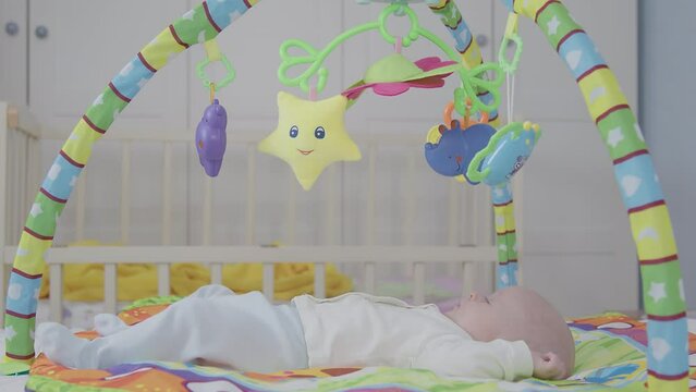 Cute newborn playmat scene, child lay on colorful mat, baby gym with engage infant, gaze at hang toy, crib background. Development activity time in nursery room, little boy look dangle plush toy.