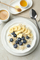 Tasty oatmeal with banana, blueberries, butter and milk served in bowl on light grey table, flat lay