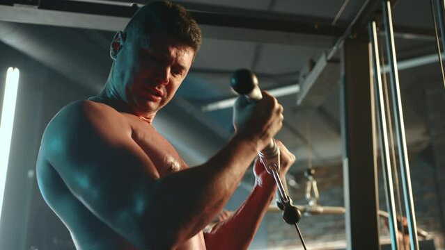 A shirtless muscular man is intensely engaged in a tricep pushdown exercise using a cable machine at a gym, displaying focus and power in his dedicated fitness routine. 