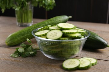 Cut cucumber in glass bowl, fresh vegetables and parsley on wooden table, closeup
