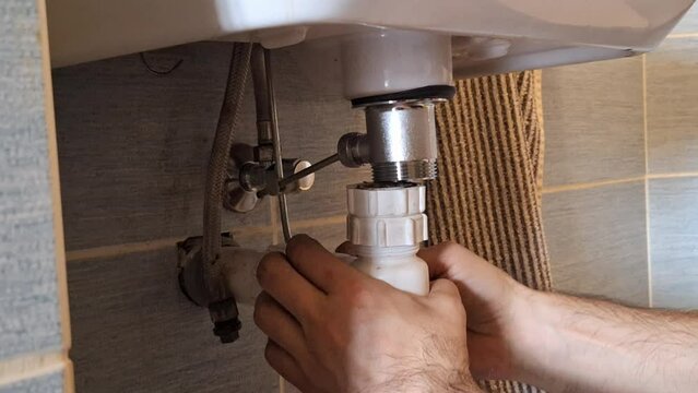 The plumber fixes the sink waste trap. A man's hands fix the installation under the washbasin in the bathroom. Concept of housekeeping, crafts, and construction.