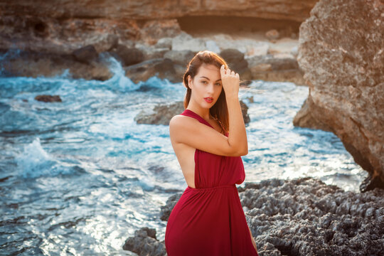 portrait of a young brunette woman standing next to the ocean shore, wearing a bordeaux red dress