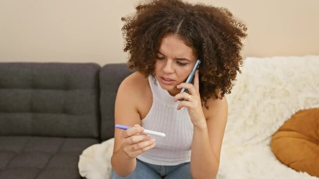 A concerned young woman with curly hair talks on the phone while holding a pregnancy test in a cozy living room. - Powered by Adobe