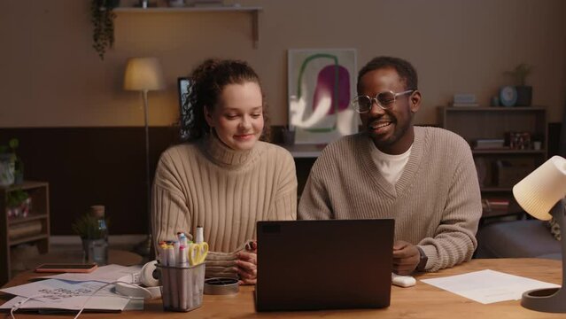 Waist up of young interracial couple sitting at desk in front of laptop and having video call at comfortable home office in afternoon