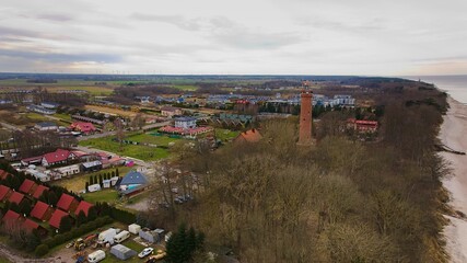 A drone captures Gąski beach, West Pomeranian Voivodeship, Poland, featuring a red brick lighthouse, Baltic Sea, sandy shore, leafless trees on dunes, holiday cottages, hotels, and residences.  © Grzegorz