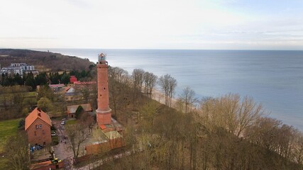 A drone captures Gąski beach, West Pomeranian Voivodeship, Poland, featuring a red brick lighthouse, Baltic Sea, sandy shore, leafless trees on dunes, holiday cottages, hotels, and residences.  © Grzegorz