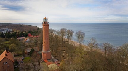 A drone captures Gąski beach, West Pomeranian Voivodeship, Poland, featuring a red brick lighthouse, Baltic Sea, sandy shore, leafless trees on dunes, holiday cottages, hotels, and residences.  © Grzegorz