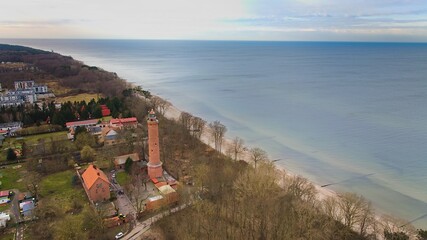 A drone captures Gąski beach, West Pomeranian Voivodeship, Poland, featuring a red brick lighthouse, Baltic Sea, sandy shore, leafless trees on dunes, holiday cottages, hotels, and residences.  © Grzegorz