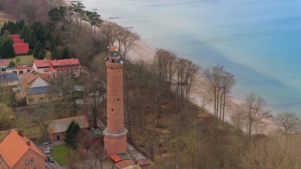 A drone captures Gąski beach, West Pomeranian Voivodeship, Poland, featuring a red brick lighthouse, Baltic Sea, sandy shore, leafless trees on dunes, holiday cottages, hotels, and residences. © Grzegorz