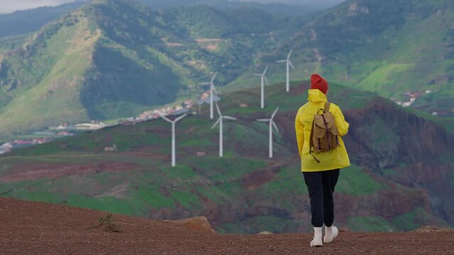 Attractive traveling woman in a yellow jacket and backpack looking at windmills in field 