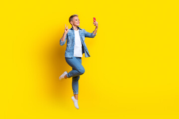 Fototapeta na wymiar Full length photo of lovely young lady jumping selfie photo show v-sign dressed stylish denim garment isolated on yellow color background