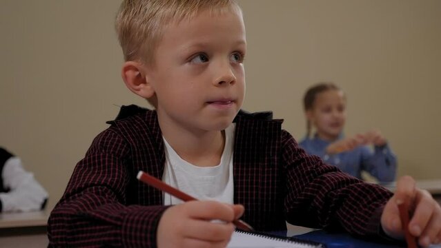 Close-up of a little boy in a shirt sitting at a desk at school, he is talking to a teacher and writing in a notebook, the learning process in elementary school.