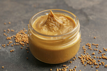 Tasty mustard sauce in jar and dry seeds on grey textured table, closeup