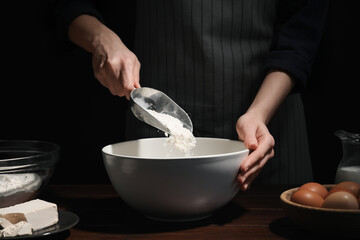 Fototapeta na wymiar Making bread. Woman adding flour into bowl at wooden table on dark background, closeup