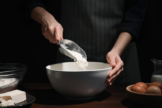 Making bread. Woman adding flour into bowl at wooden table on dark background, closeup