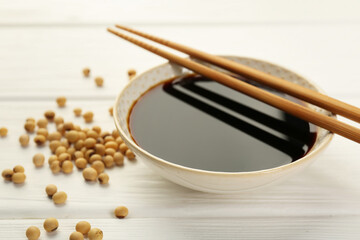 Tasty soy sauce in bowl, chopsticks and soybeans on white wooden table, closeup