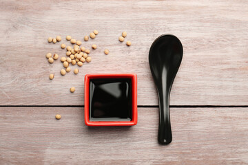 Soy sauce in bowl, soybeans and spoon on wooden table, flat lay