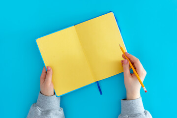 Open yellow notepad in female hands on a blue background, top view.