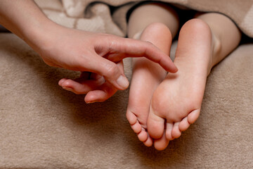 Children's feet under a blanket are tickled by their mother's hand - obrazy, fototapety, plakaty