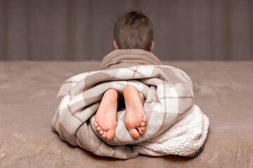 The boy's legs, covered with a blanket, lie on the bed in the bedroom.