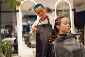 Stylish female hairdresser applying spray on wet hair to woman in beauty salon