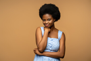 Calm black female with shy sincere smile, natural curly hair, wearing blue summer dress studio blank. Pleased african american woman looking to camera, isolated on beige background for advertisement.