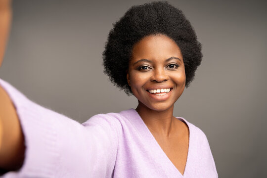 Charismatic Optimistic African American Female With Outstretched Making Selfie Shot Smiling Broadly With White Teeth, Looking At Camera. Closeup Face Glad Happy Black Woman Isolated On Grey Background