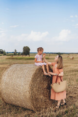 Family mother and daughter in a field of mown wheat against the blue sky. Little daughter sits on a bale of straw, mother touches her daughter's face with her hand