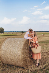 Mom and little daughter in a field of harvested wheat against the blue sky. Mom is holding a wicker linen bag in her hands. Daughter kisses mom