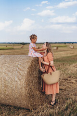 Naklejka na ściany i meble Mom and little daughter in a field of harvested wheat against the blue sky. Mom is holding a wicker linen bag in her hands. Daughter touches her mother's face with her hand