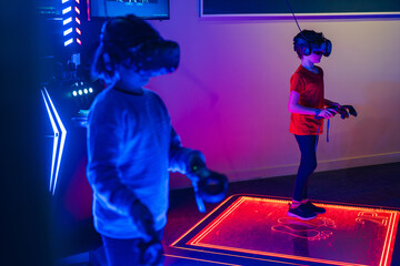 Two anonymous kid with VR headsets are playing on an interactive, illuminated gaming platform in a neon-lit room