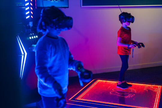 Two anonymous kid with VR headsets are playing on an interactive, illuminated gaming platform in a neon-lit room