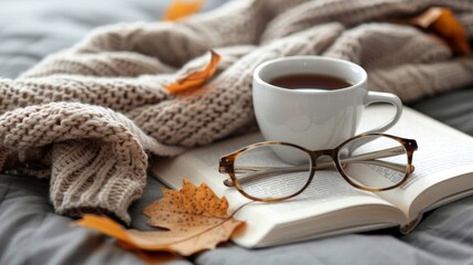 Cozy hygge concept with beige sweater, tea or coffee mug, book, and glasses on gray bed.