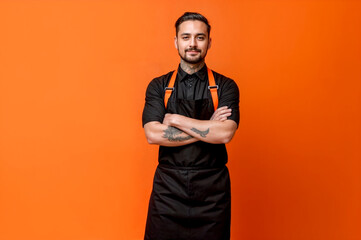 A young man, a barista, a bartender in a black apron, wearing a white T-shirt, keeps his arms crossed and stands isolated on orange studio background.