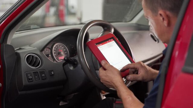 Auto electrician uses OBD2 scanner and tablet for car diagnostics. Mechanic checks vehicle engine codes in workshop. Professional tech analysis, fix auto problems. Dashboard, tools in use.