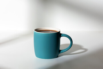 A cup of coffee stands on a white background with a shadow from the window in the morning light