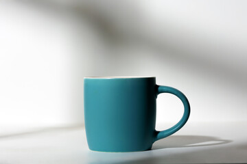 A cup of coffee stands on a white background with a shadow from the window in the morning light