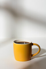 A cup of coffee stands on a white background with a shadow from the window in the morning light