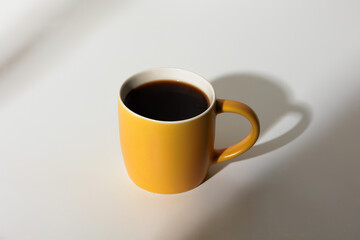 A cup of coffee stands on a white background with a shadow from the window in the morning light