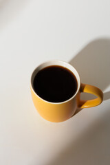 A cup of coffee stands on a white background with a shadow from the window in the morning light