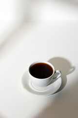A cup of coffee stands on a white background with a shadow from the window in the morning light