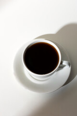 A cup of coffee stands on a white background with a shadow from the window in the morning light