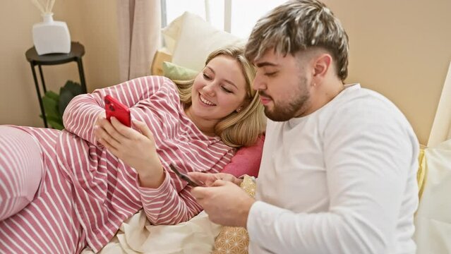 A smiling woman and man cuddled in bed with a smartphone, depicting love and comfort in a cozy bedroom setting.