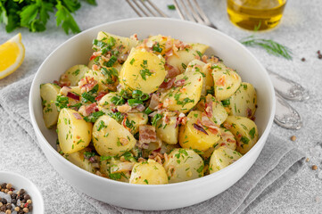 Warm potato salad with bacon, onion, fresh herbs and vinegar dressing in a bowl on a gray concrete background. German Kartoffelsalat.