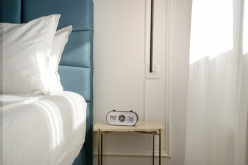 Blue king size bed with crisp white bedding and digital alarm clock on the nightstand under beautiful morning sunlights